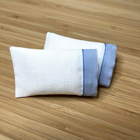 Two white cotton dollhouse miniature pillowcases with blue trim 1:12 scale, laid flat on a wooden surface